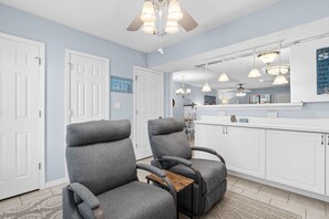 A room with two grey armchairs, a white ceiling fan, and a kitchen area with white cabinets and pendant lights. - Sunshine & Shenanigans- Steps to Main Pool & More! (Panama City Beach)