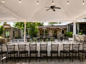 An outdoor dining area with tables set for six, surrounded by stone walls and a ceiling fan.