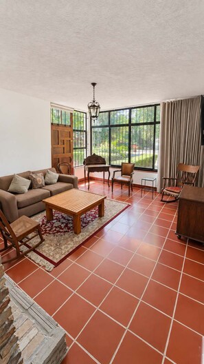 Amplia sala de estar con sofá, mesa de centro y sillas, ventanas grandes con cortinas y vistas a la vegetación exterior.