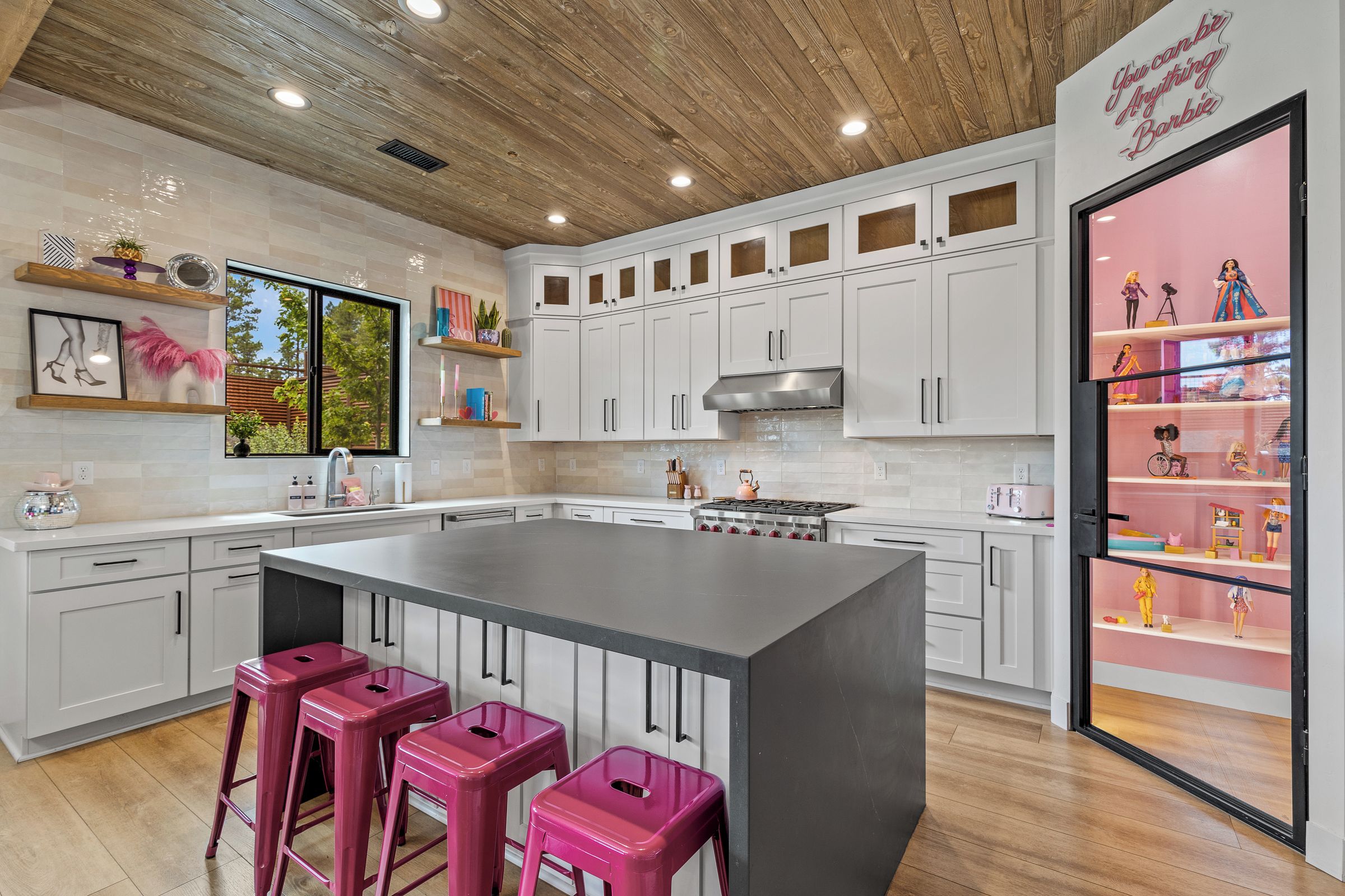 A modern kitchen with white cabinets, a large island with pink stools, and a decorative shelf with figurines.