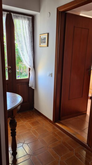 Una habitación con puerta de madera, ventana con cortinas de encaje, un cuadro en la pared y una mesa redonda.