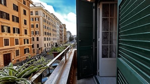 Ein Balkon mit Blumentöpfen, Blick auf eine Straße mit abgestellten Motorrädern und ein Gebäude mit mehreren Fenstern und Fensterläden.