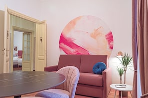 Ein Zimmer mit einem runden Tisch, einer Couch, einer Wand mit einem großen abstrakten Gemälde und einem kleinen Beistelltisch mit einer Pflanze.