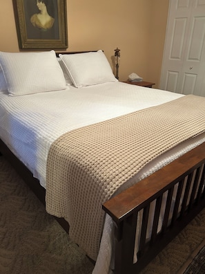 Una cama con colcha blanca y manta beige, una mesita de noche con lámpara y un cuadro enmarcado en la pared.