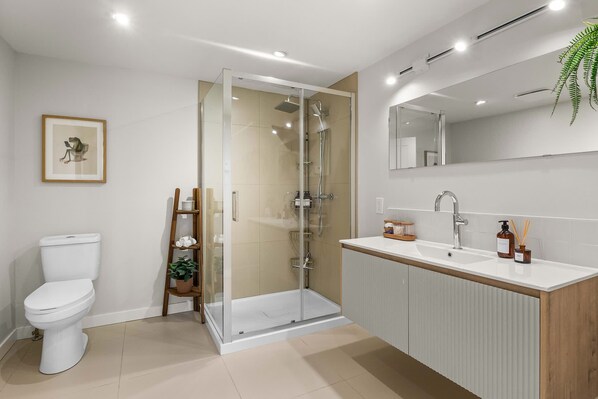Un baño moderno con ducha envuelta en vidrio, inodoro blanco y tocador de madera con espejo.