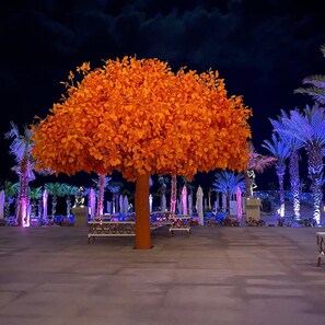 Un gran árbol artificial con follaje anaranjado en un espacio al aire libre con palmeras y bancos.