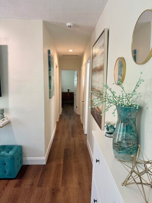 A hallway with wooden flooring, white walls, a teal ottoman, a white dresser with drawers, a vase with green branches, and two mirrors. - Newly renovated custom 3-bedroom/ 2 bath house in coastal area! (West Palm Beach)