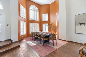 A dining area with a round table set for six, surrounded by chairs, in a room with orange walls and large windows. - Mesa Resort • 6BR • Pool + Slide • Spa • Pickleball • Theater (Mesa)