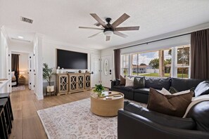 A modern living room with a large flat-screen TV, a black leather sofa, a round coffee table, and a ceiling fan. - St Pete Poolside Paradise w/ Outdoor Bar (St. Petersburg)
