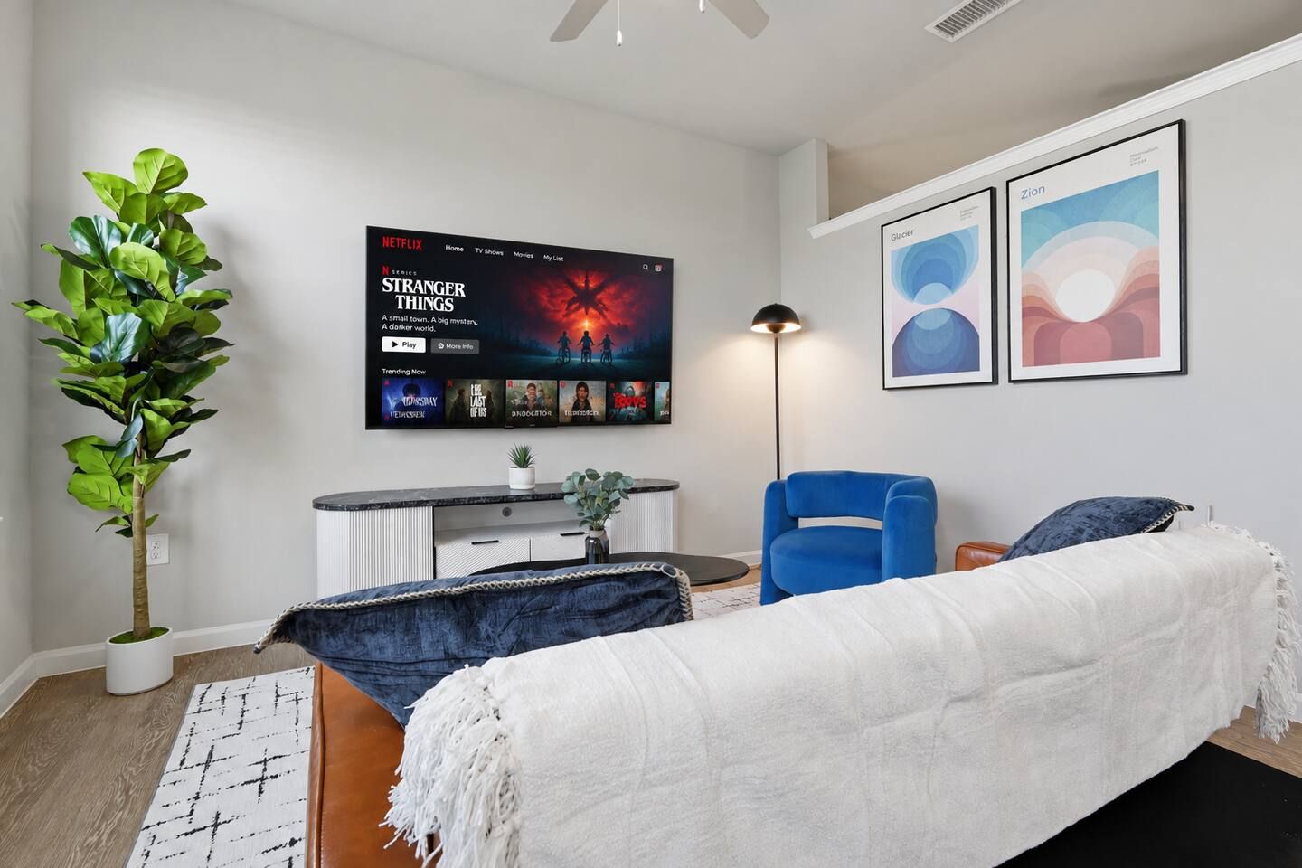 A modern bedroom with a bed, a blue chair, a TV displaying 'Stranger Things', and two framed artworks on the wall.