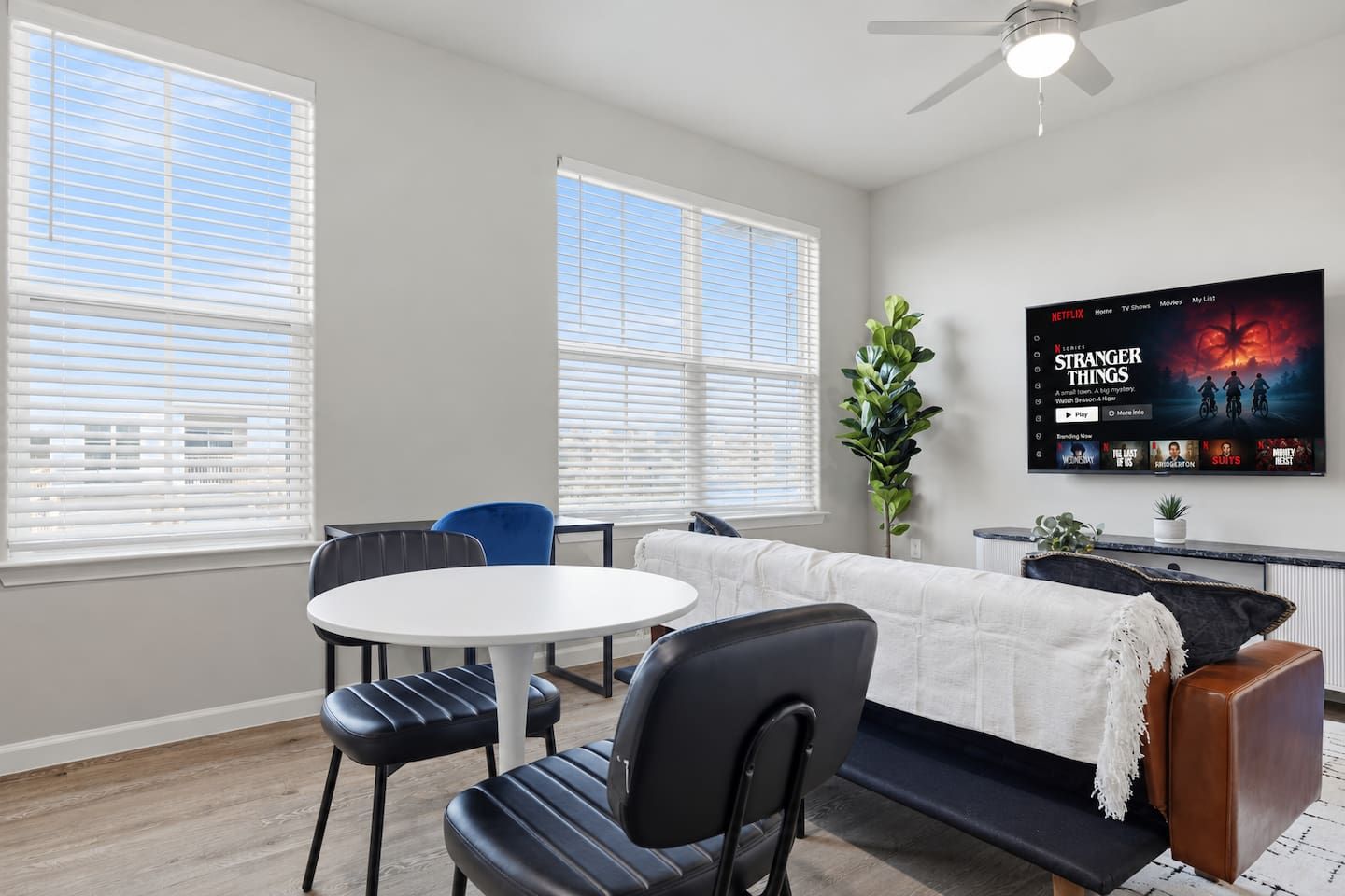 A modern living room with a flat-screen TV displaying a movie, a round dining table, two chairs, a sofa, and a ceiling fan.