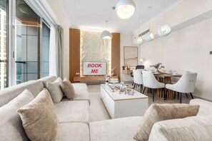 A modern living room with a sofa, coffee table, dining area with chairs, and a 'BOOK ME' sign. - Stayshort - Elegant 2 Bedroom That Sleeps 4 (Dubai)