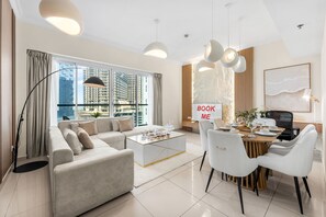 A modern living room with a sofa, coffee table, dining table, and chairs. There is a sign that says 'BOOK ME' on the wall. - Stayshort - Elegant 2 Bedroom That Sleeps 4 (Dubai)