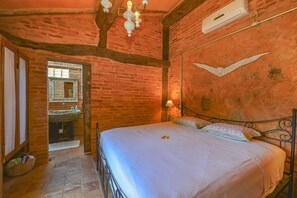 Ein rustikales Schlafzimmer mit freiliegenden Backsteinwänden, einer Holzbalkendecke, einem Bett mit weißen Leinen, einer Pendelleuchte und einer Wand-Klimaanlage.