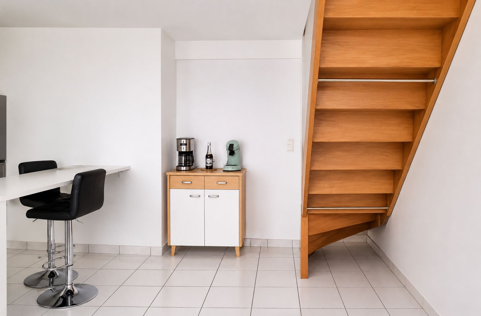 Een moderne keuken met een wit aanrecht, twee zwarte barkrukken, een houten kast met lades, een koffiezetapparaat en een groene waterkoker.