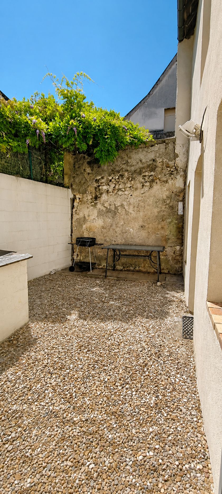Une cour avec un sol en gravier, un banc, un barbecue et un mur recouvert de plantes grimpantes.