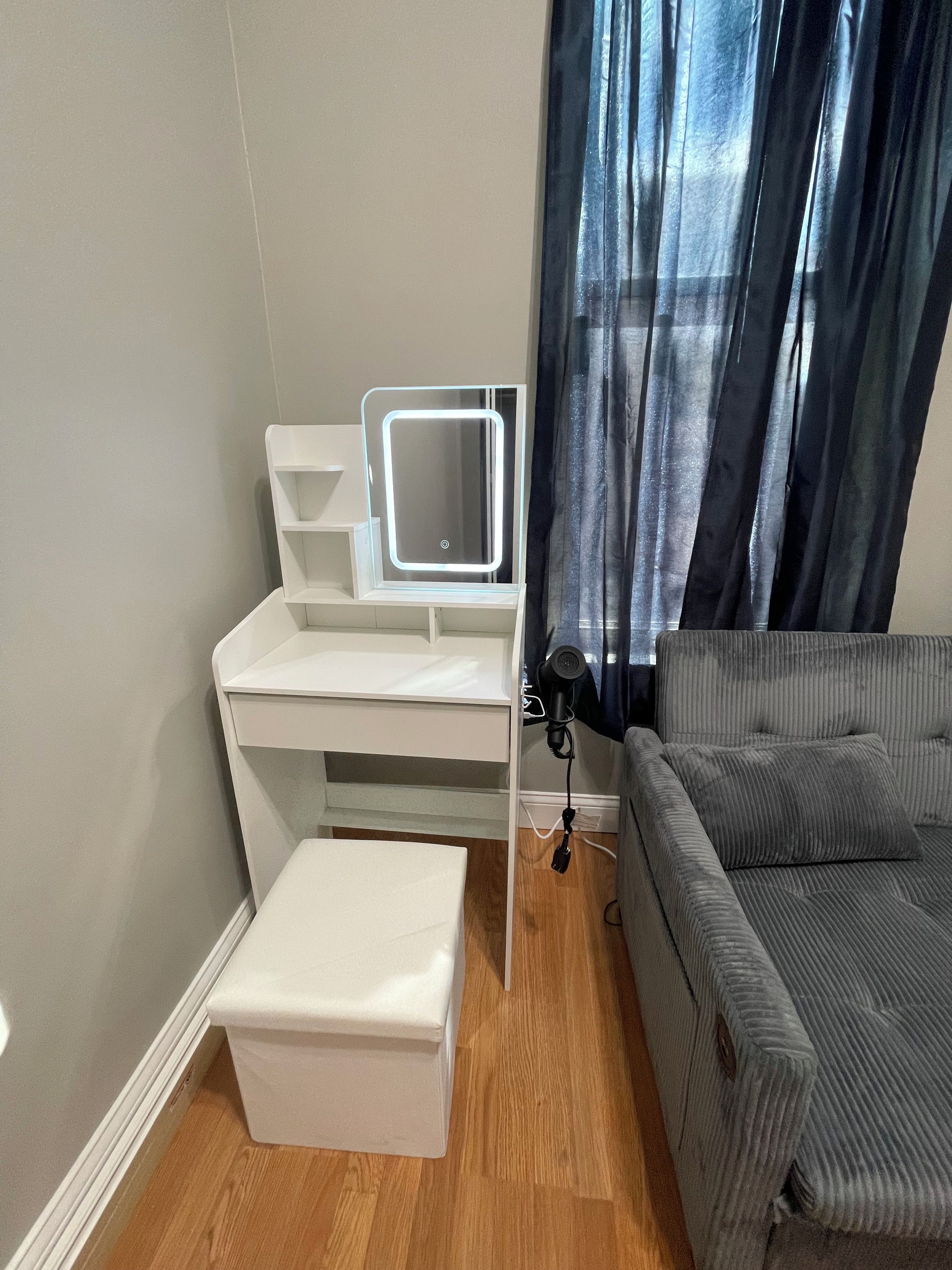 A room with a white desk, a mirror, a grey sofa, and a small white ottoman.