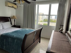 Una habitación de hotel con una cama, una mesita de noche, una lámpara, una ventana con cortinas y vistas a edificios.