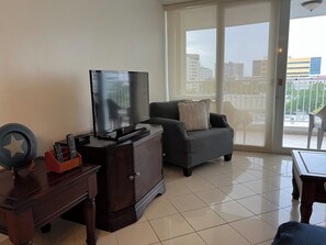 A living room with a flat-screen TV on a wooden cabinet, a sofa with a cushion, a coffee table, and a view of a cityscape through large windows. - Beautiful La Mancha Apartment + free parking (Carolina)