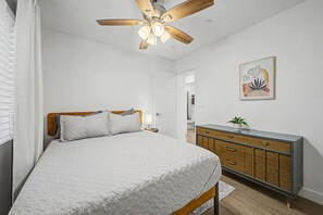 Ein Schlafzimmer mit einem Bett, einem Deckenventilator, einem Schminktisch und einer Tür zu einem anderen Raum.