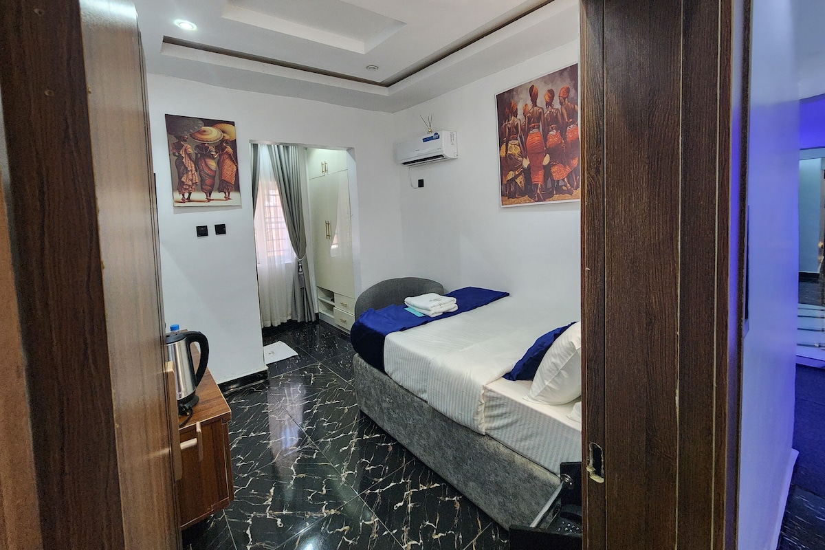 A hotel room with a bed, a wardrobe, a desk with a kettle, and a painting on the wall.