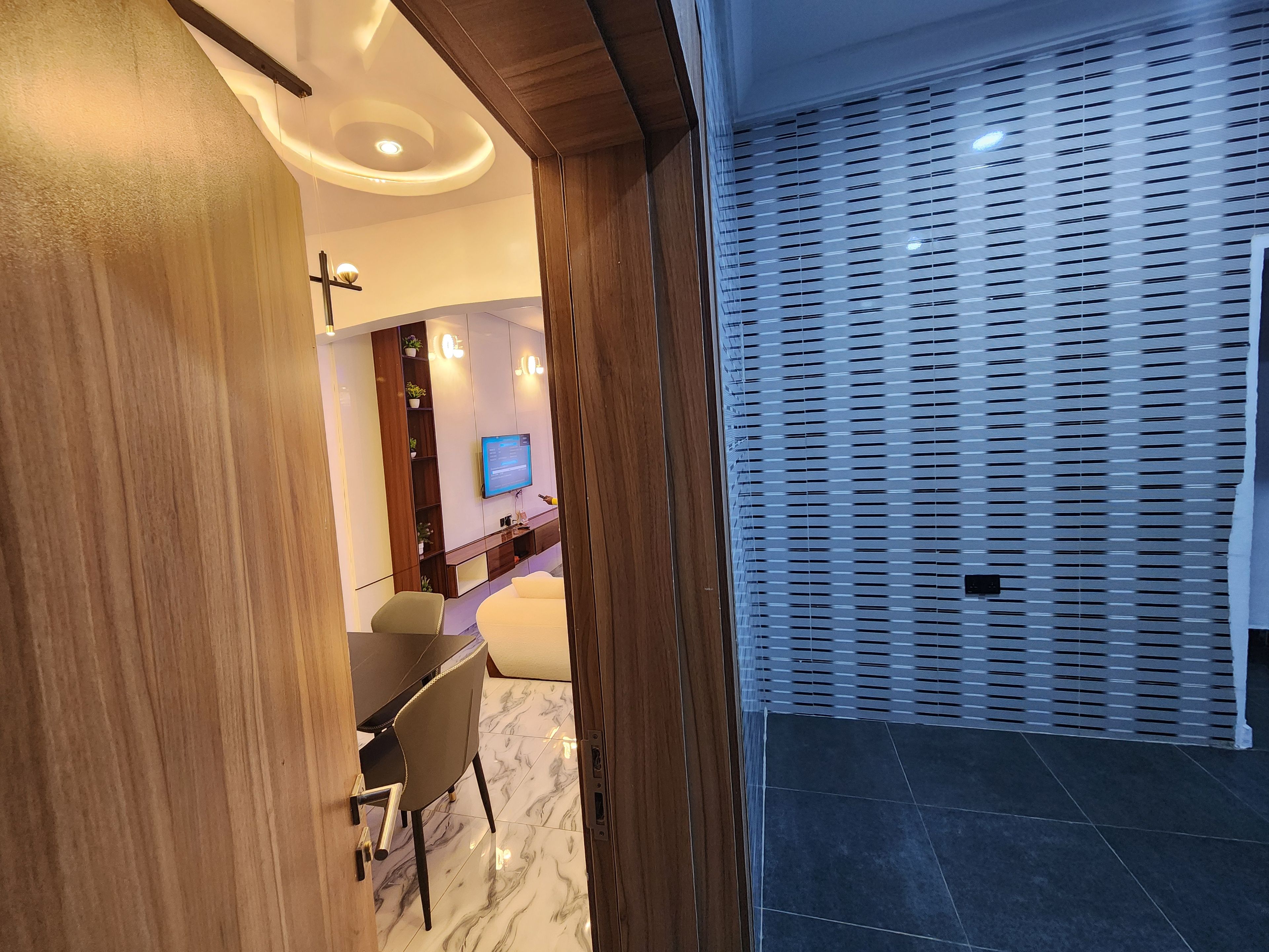 Un interior moderno con una puerta de madera, una zona de comedor con mesa y sillas, un televisor montado en la pared y un papel tapiz decorativo.