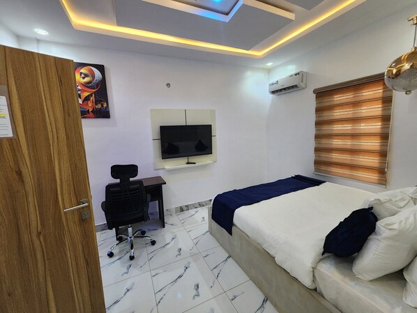A hotel room with a bed, a desk with a chair, a television mounted on the wall, and an air conditioning unit.