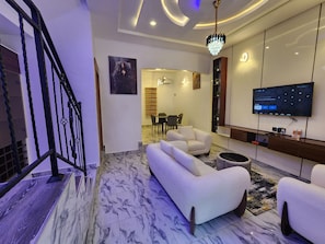 A modern living room with a marble floor, a staircase, a flat-screen TV, and a dining area.