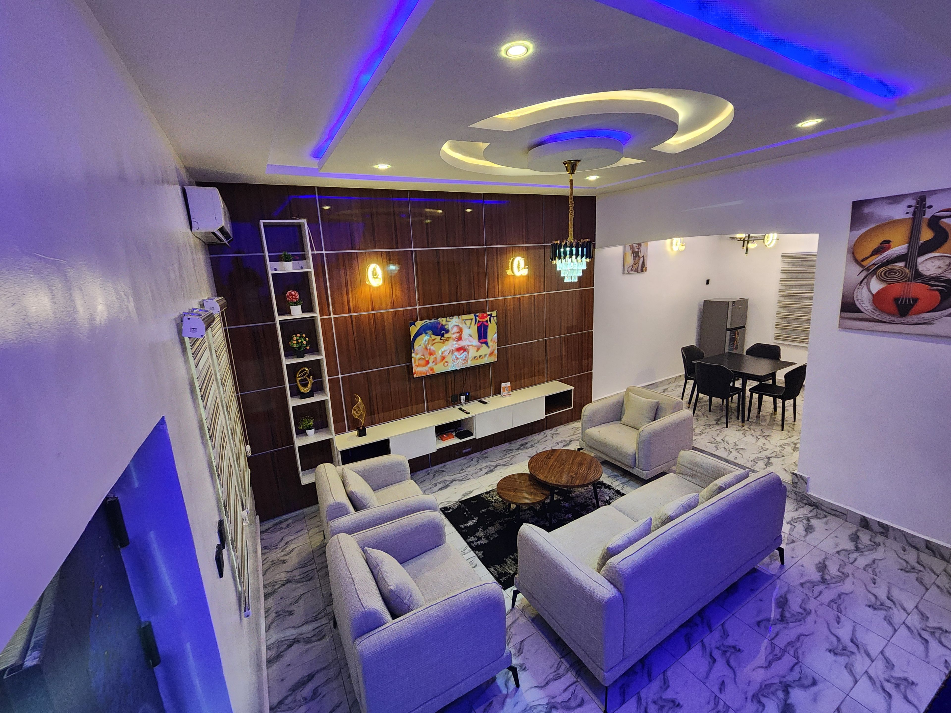 A modern living room with a sofa set, a TV mounted on the wall, and a dining area with chairs and a table.