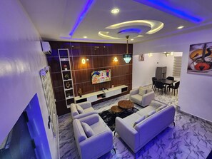 A modern living room with a sofa set, a TV mounted on the wall, and a dining area with chairs and a table.