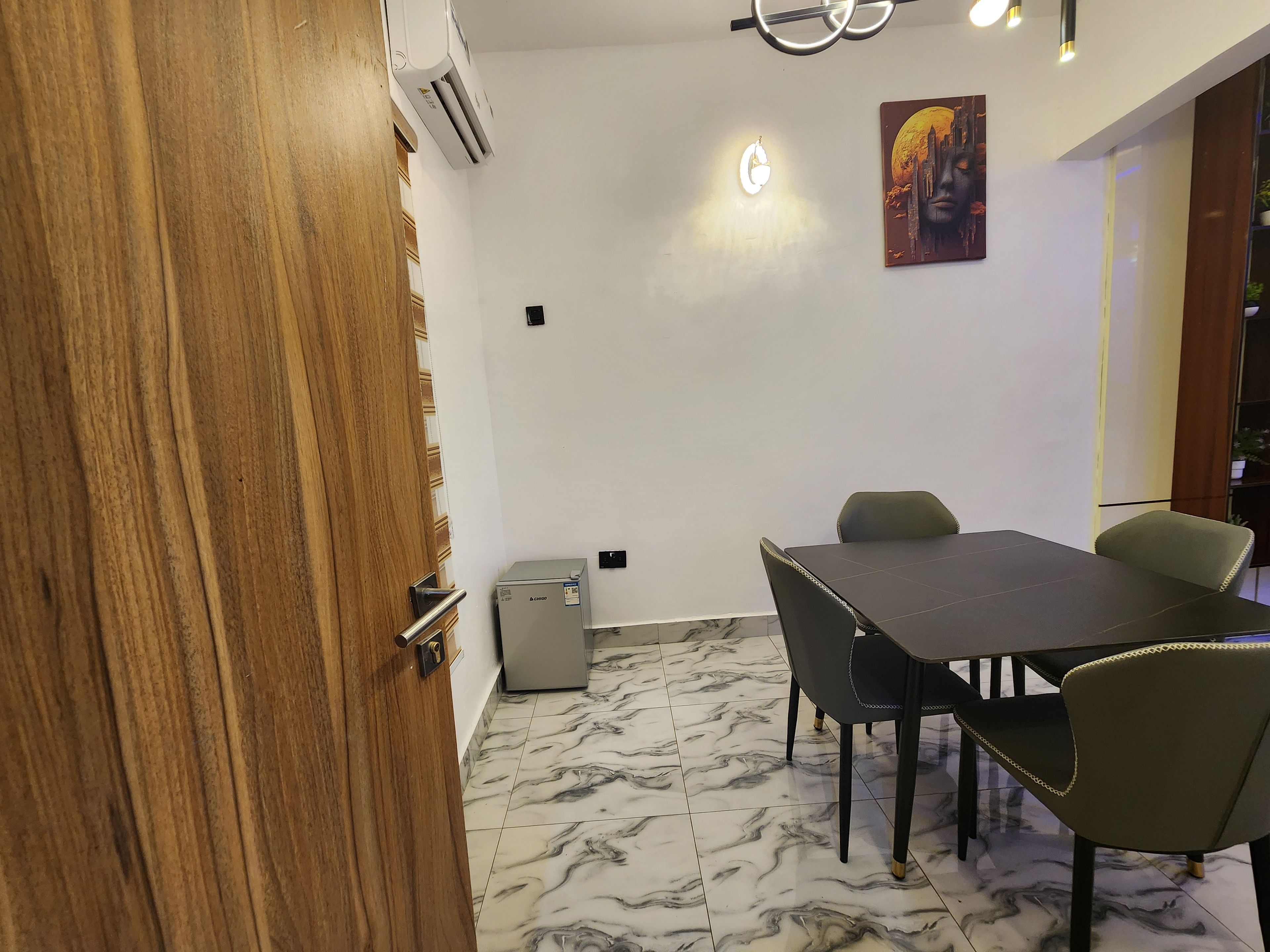 A room with a wooden door, a table with chairs, a wall-mounted air conditioner, a light fixture, and a painting.
