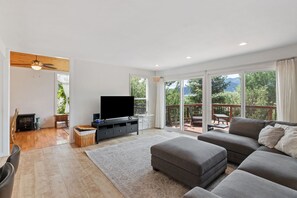 En moderne stue med et fladskærms-tv, en grå sofa og en balkon med udsigt over grønne områder.