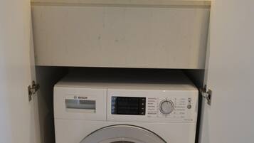 A Bosch washing machine in a compact cabinet with a shelf above it containing decorative items.