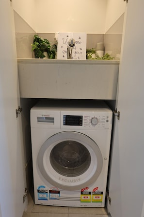 Un lave-linge Bosch dans un meuble compact, avec une étagère au-dessus contenant des objets décoratifs.