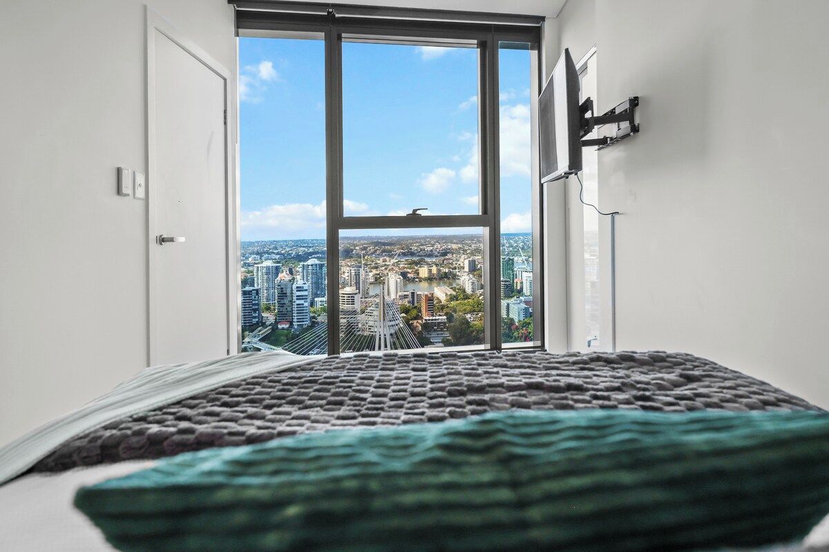 A room with a large window, a bed with a patterned bedspread, and a wall-mounted TV.
