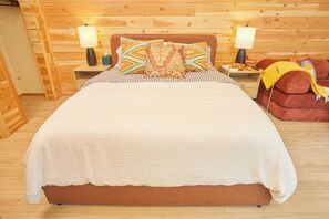 Ein Bett mit weißer Bettdecke und gemusterten Kissen, flankiert von Nachttischen mit Lampen und ein roter Sessel mit gelber Decke.