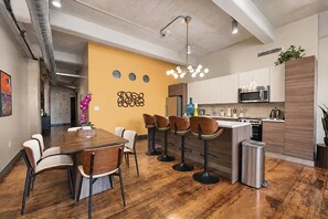 A modern kitchen with a central island, bar stools, and a dining area with a wooden table and chairs. - The Bentley Collection - The Marquee (Dallas)