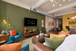 A modern living room with a flat-screen TV mounted on the wall, a dining area with a wooden table and chairs, and a kitchen area with a yellow wall and decorative lights. - The Bentley Collection - The Marquee (Dallas)