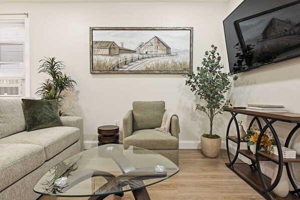 Een woonkamer met een bank, fauteuil, glazen salontafel, potplanten en een schilderij aan de muur.