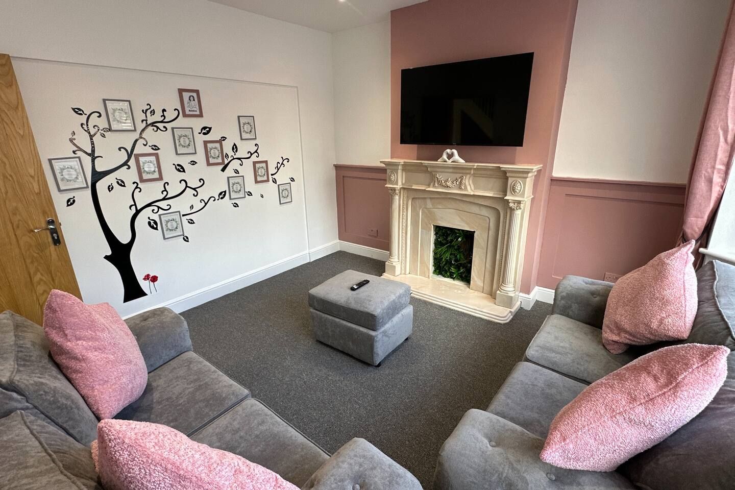 A living room with a fireplace, grey sofas, a flat-screen TV, and a wall with a tree decal.