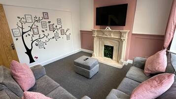 A living room with a fireplace, grey sofas, a flat-screen TV, and a wall with a tree decal.