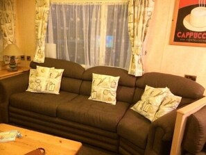 A living room with a sofa, pillows, a table, and curtains. - Seashore Holiday Park - Holiday Accommodation 5432 (Great Yarmouth)