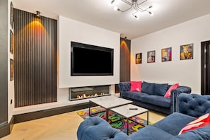A modern living room with a flat-screen TV, a fireplace, a blue sofa, and colorful artwork on the walls. - 1 Bedford House - Close to Town and Seafront (Tenby)