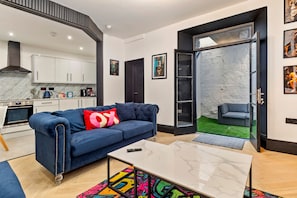 A modern living room with a blue sofa, a coffee table, and a kitchen in the background. - 1 Bedford House - Close to Town and Seafront (Tenby)