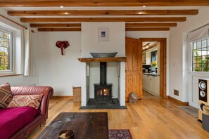 Ein gemütliches Wohnzimmer mit Kamin, Holzbalken, einer Couch und einer durchgehenden Tür erkennbaren Küche.