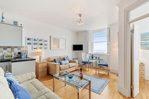 Ein modernes Wohnzimmer mit einer Couch, einem Couchtisch und einem Essbereich mit Stühlen und einem Tisch. Ein Fernseher ist an der Wand montiert und es gibt ein Fenster mit Rollläden.