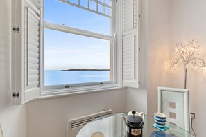 Ein Zimmer mit Meerblick durch ein Fenster mit weißen Fensterläden, ein Glastisch mit Kaffeekanne und Tasse sowie eine Zierpflanze.
