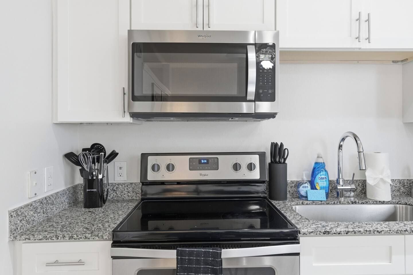 A kitchen with a stainless steel microwave, a black stove, white cabinets, a granite countertop, a sink with a faucet, and a dish soap bottle.
