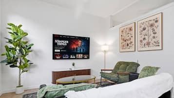 A living room with a flat-screen TV displaying 'Stranger Things', a green armchair, a white sofa, a small round coffee table, and a potted plant.