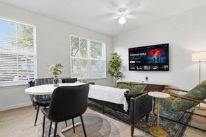A modern living room with a dining area, a flat-screen TV mounted on the wall, and a ceiling fan. - The Austin Haven | Stylish 1Bed + Pool&Gym (Pflugerville)
